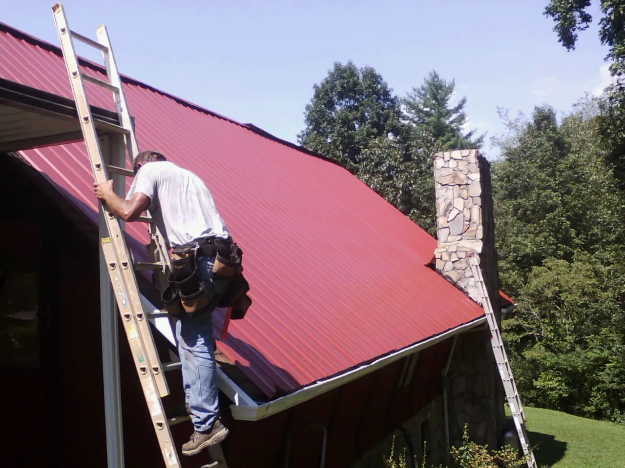 Roofer on a ladder beside a red metal roof and stone chimney — Soffit & Fascia Repair by Roofing Brothers in Swansea